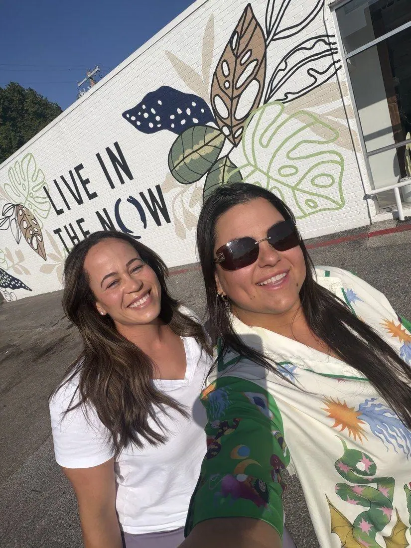 Two smiling creators taking a selfie in front of the Live in the NOW mural