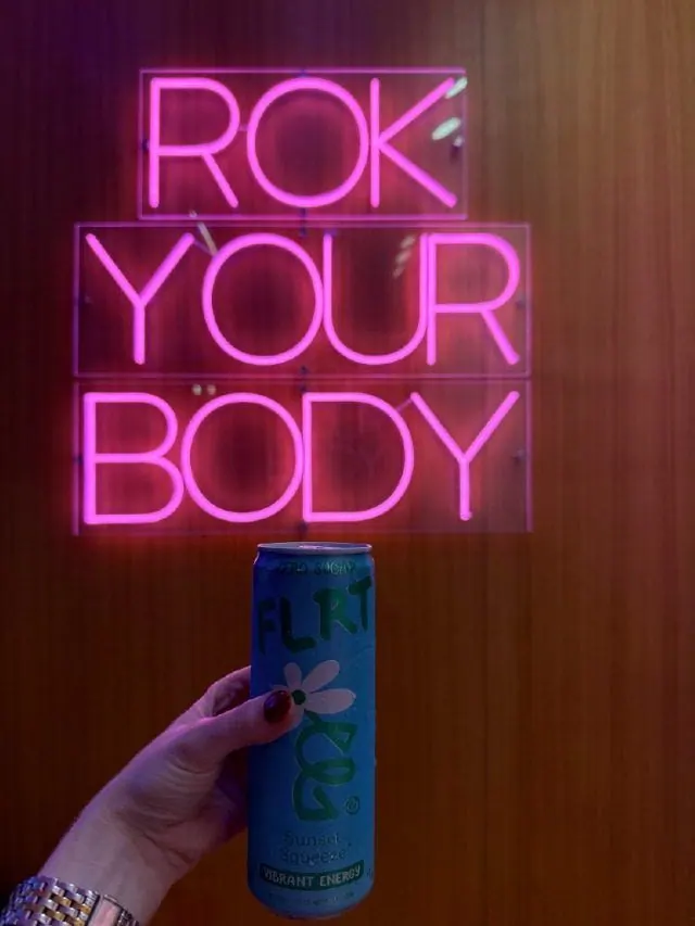Hand holding a drink can beneath a pink ROK YOUR BODY neon sign