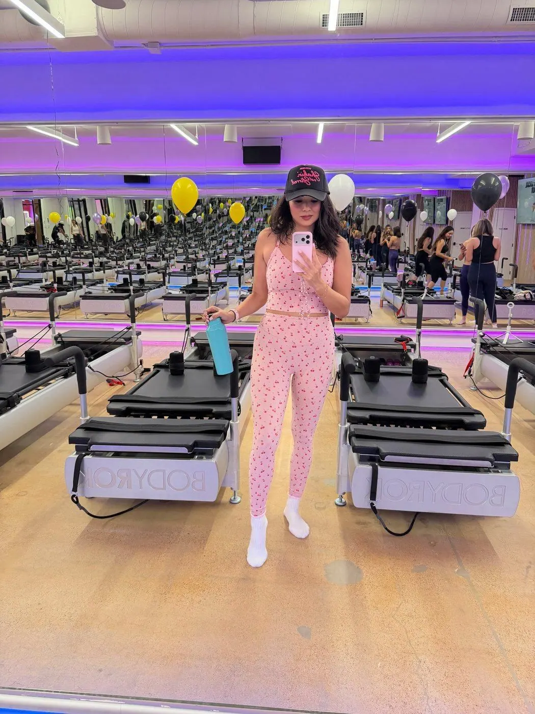 Creator in a cherry-print set taking a mirror selfie between reformers, balloons behind her