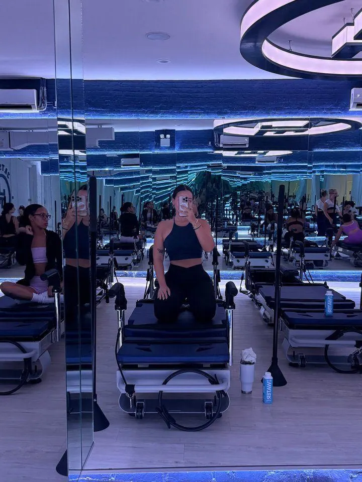 Creator kneeling on a reformer for a mirror selfie in a busy blue-lit pilates class