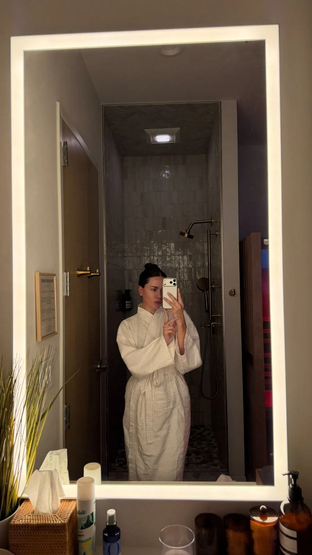 Creator in a white robe taking a selfie in a lit spa bathroom mirror