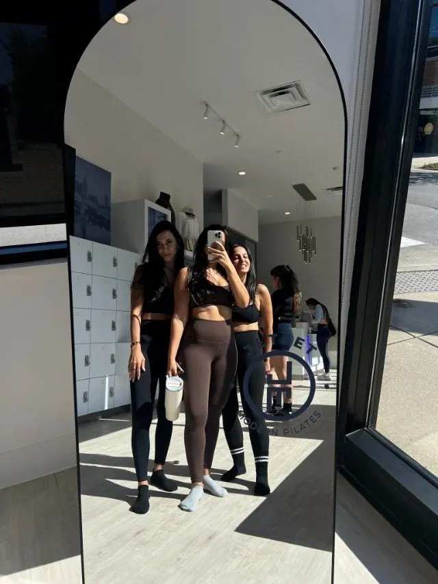 Three friends in workout clothes taking a mirror selfie in a pilates studio lobby