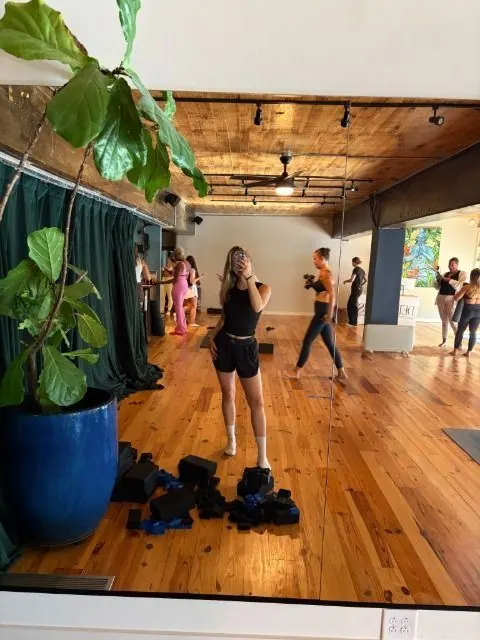 Creator taking a mirror selfie in a sunny studio with wood floors as others work out